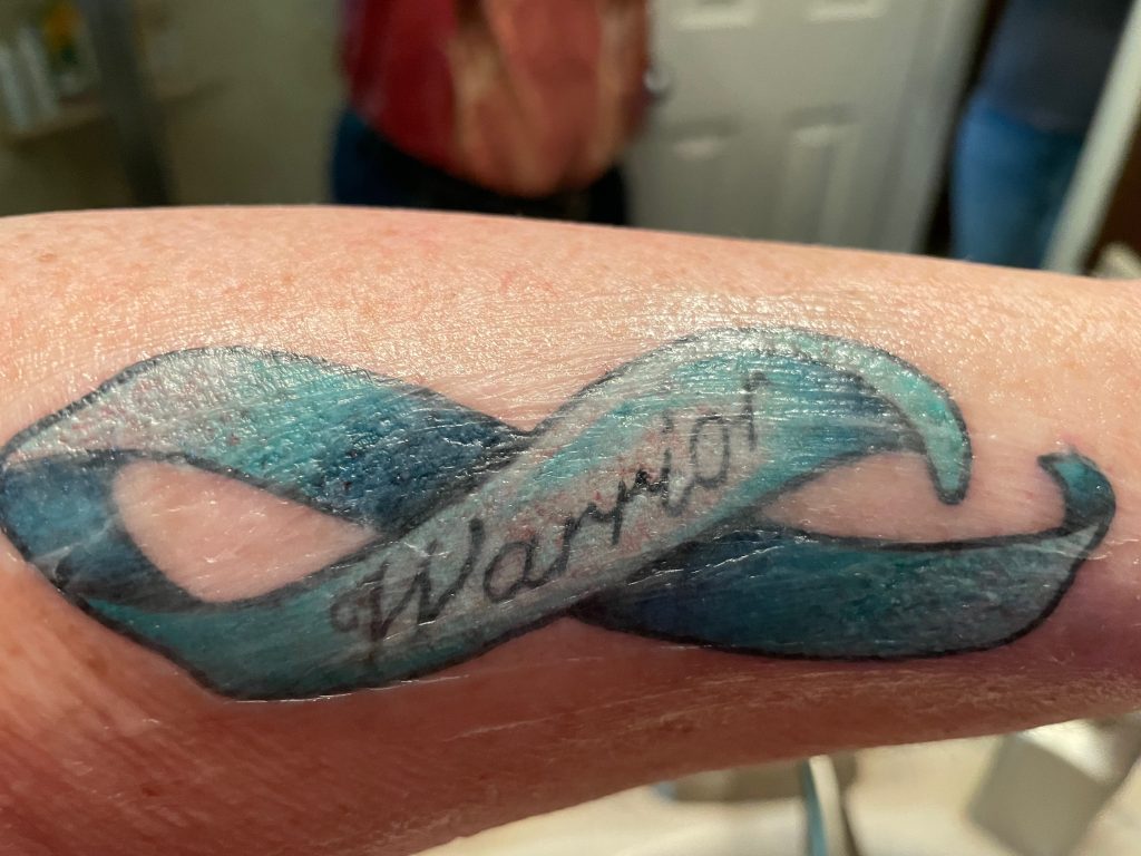 teal ribbon tattoo with the word warrior in script.